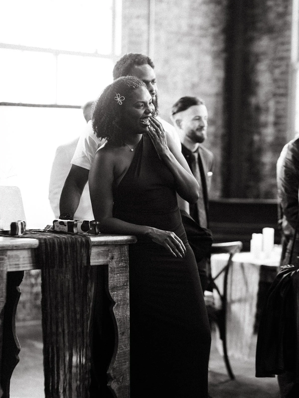 Wedding guest smiles and reacts during a reception speech while leaning against a rustic wooden table inside an industrial wedding venue with exposed brick walls.