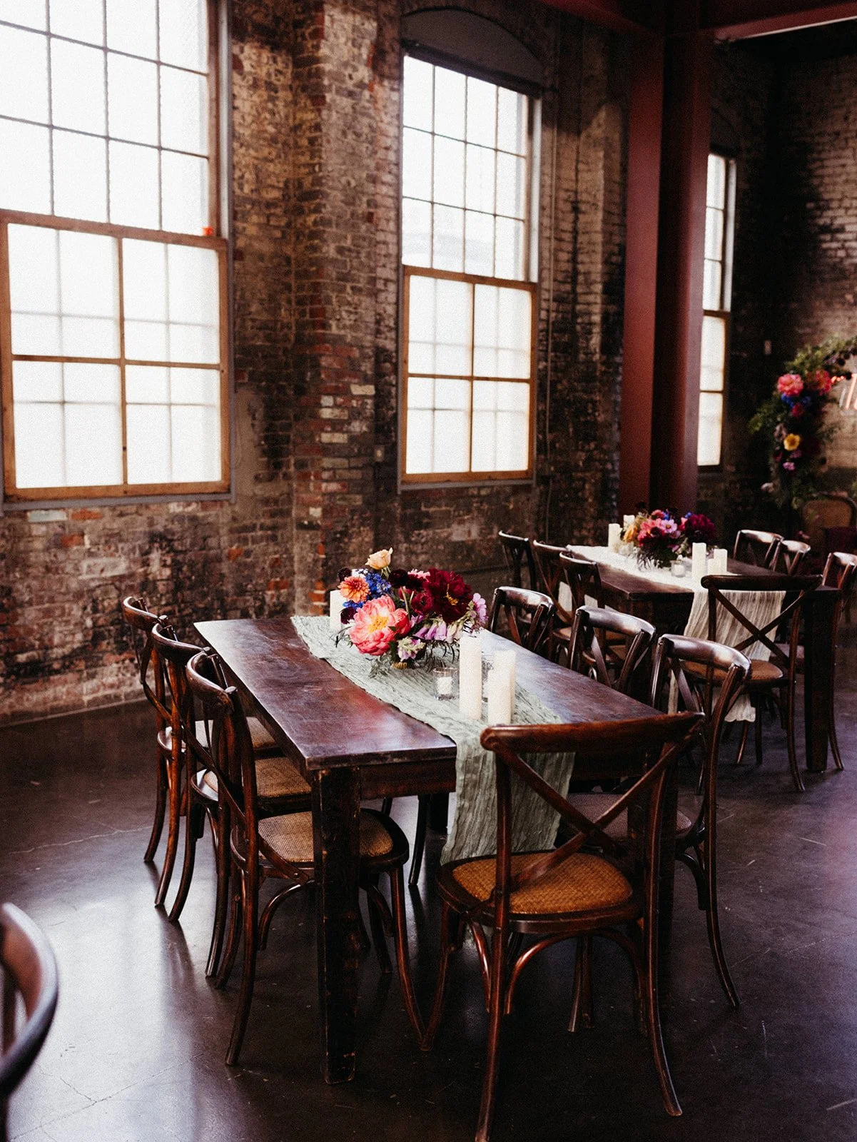 Long wooden reception tables with colorful floral centerpieces and candles inside an industrial wedding venue with exposed brick walls and large windows.