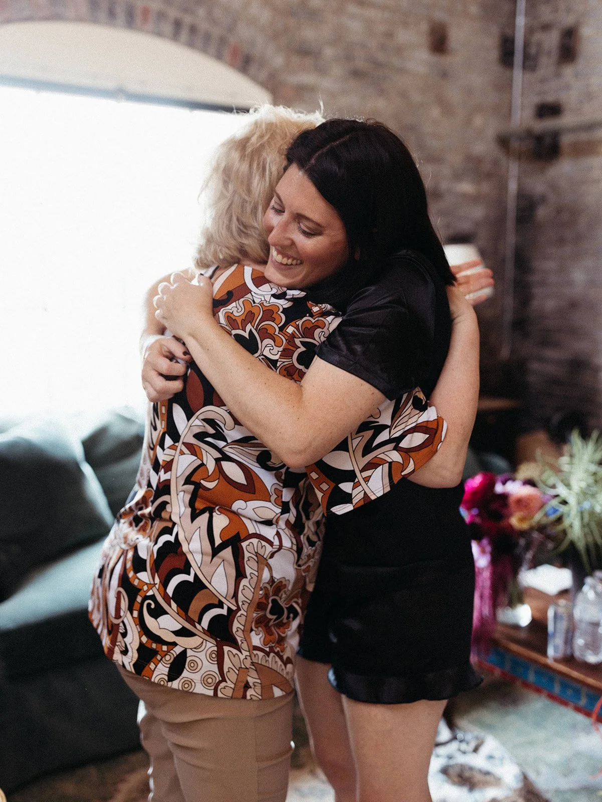 Bride wearing black satin getting-ready pajamas hugs a loved one inside an industrial loft wedding venue before the ceremony.