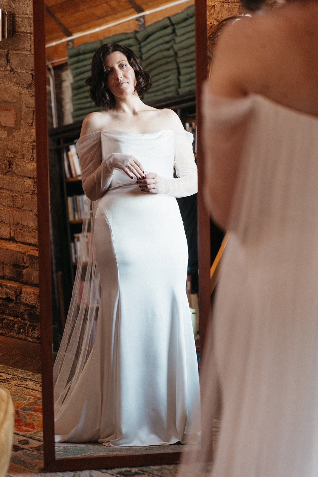 Bride in an off-the-shoulder satin wedding gown looks at her reflection in a full-length mirror while getting ready in a historic loft venue.
