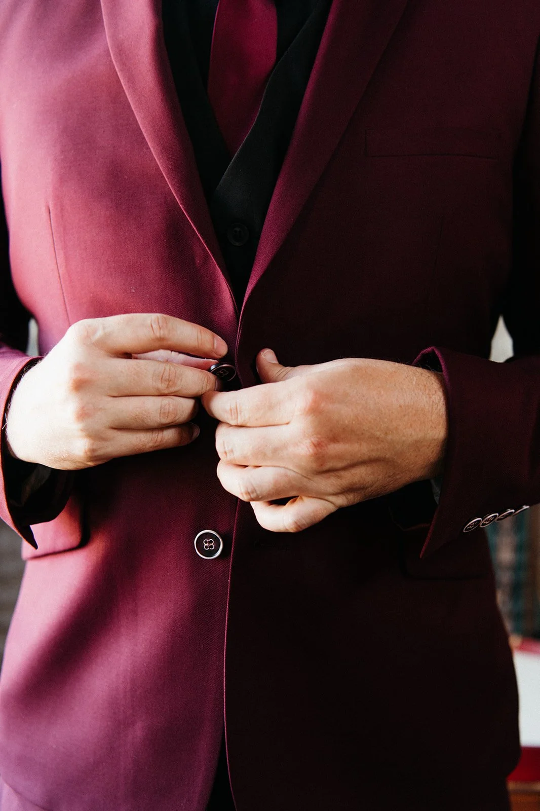 Close-up detail of the groom fastening the button on a burgundy wedding suit jacket during wedding preparations.