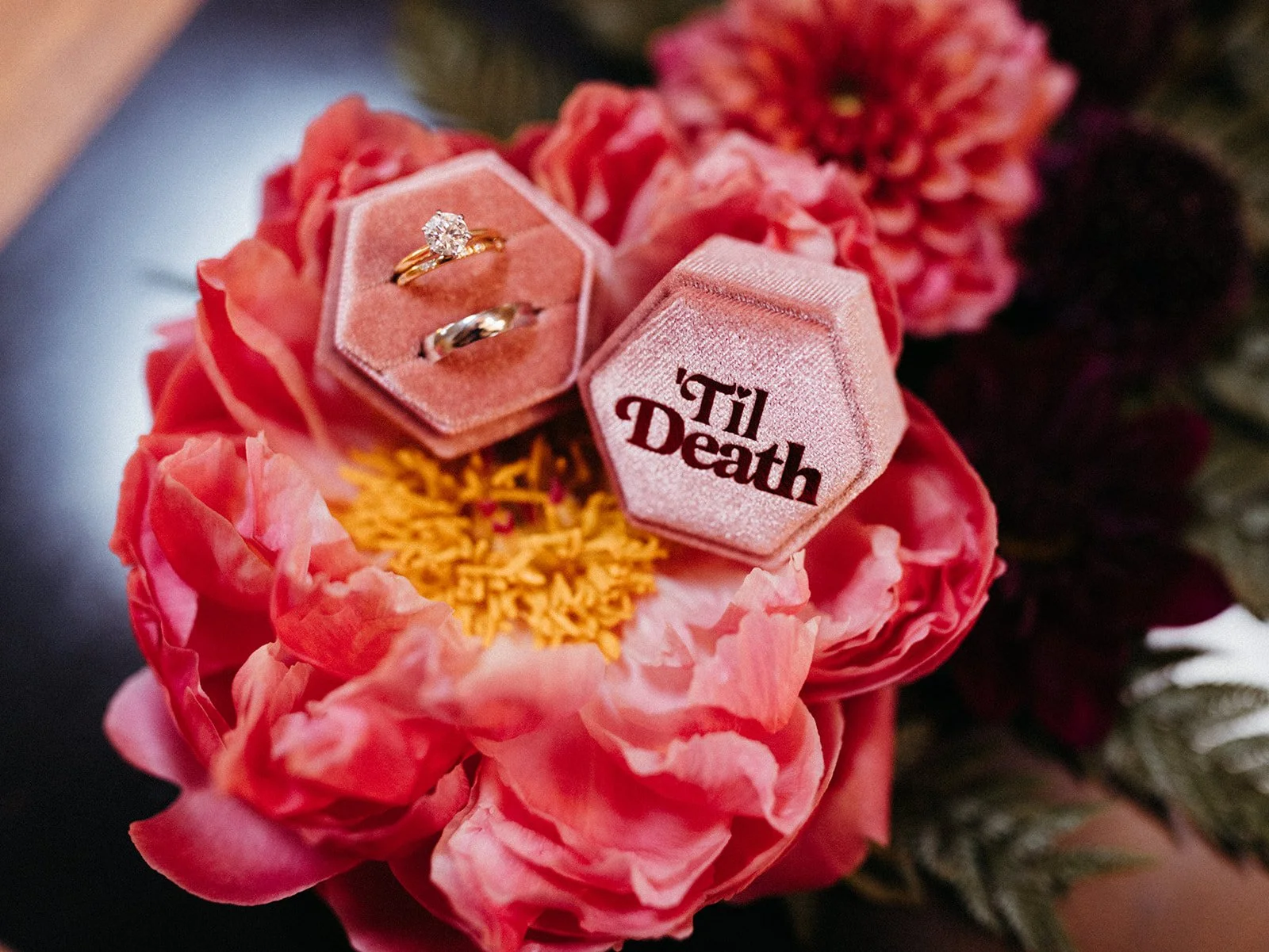 Wedding bands and engagement ring displayed in pink velvet ring boxes with "'Til Death" lettering atop a vibrant pink flower arrangement.