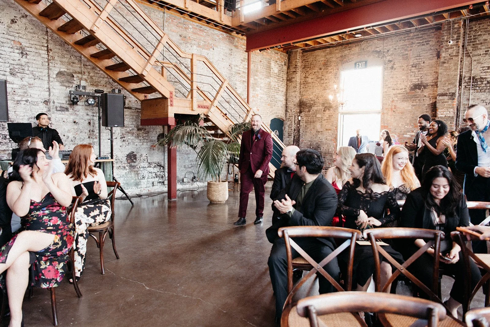 Groom in a burgundy suit walks down the aisle as wedding guests applaud during an intimate industrial loft ceremony.