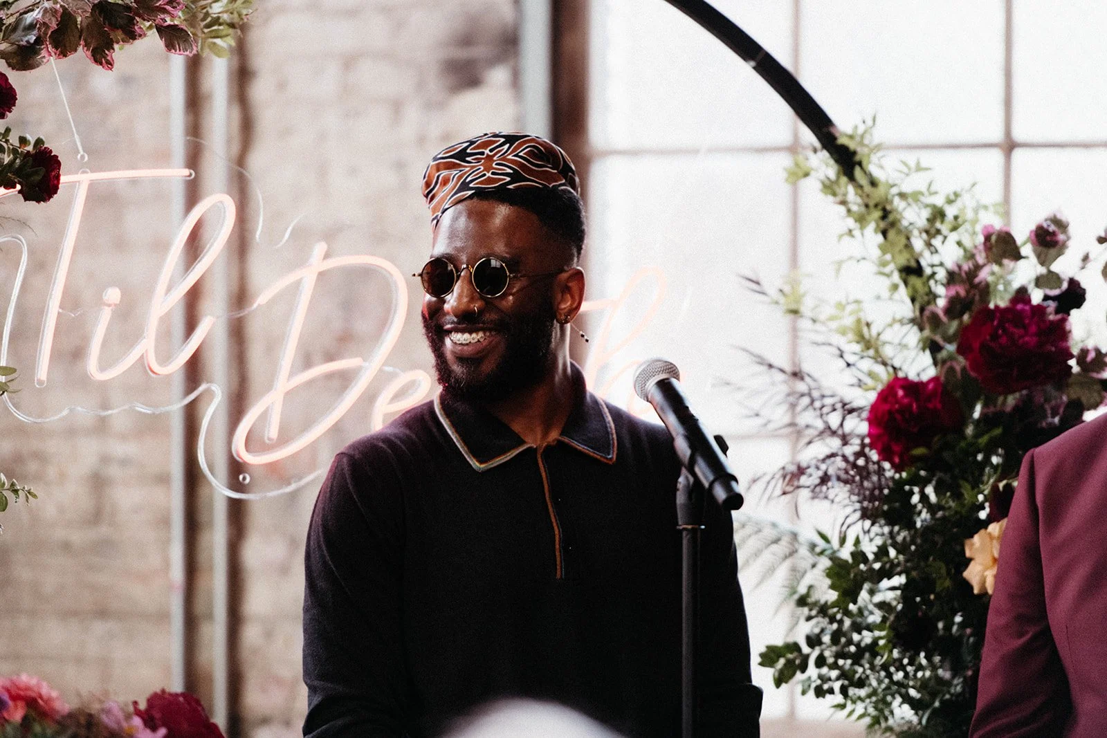 Wedding officiant smiles beside a microphone during a ceremony framed by colorful floral arrangements and a neon sign backdrop.