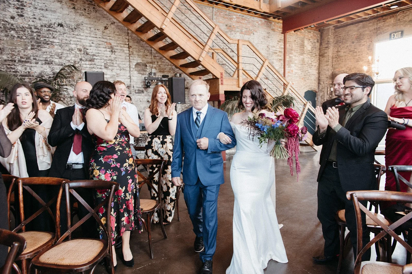 Bride carrying a colorful bouquet walks down the aisle with a family member as guests stand and applaud during the wedding ceremony.