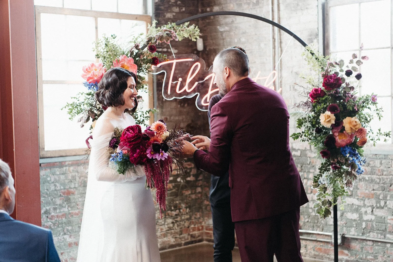 Bride and groom exchange rings beneath a floral ceremony arch with a neon sign backdrop during an industrial loft wedding ceremony.