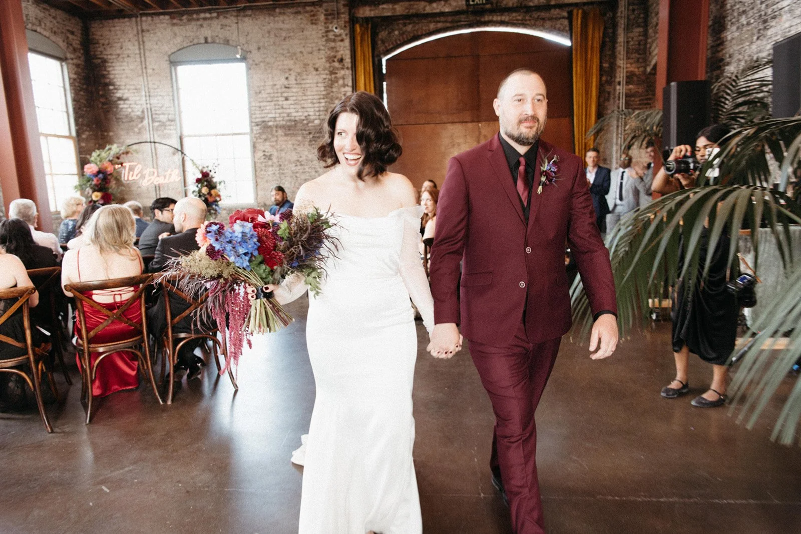 Newlyweds walk down the ceremony aisle smiling and holding hands inside a historic industrial venue surrounded by guests and reception décor.