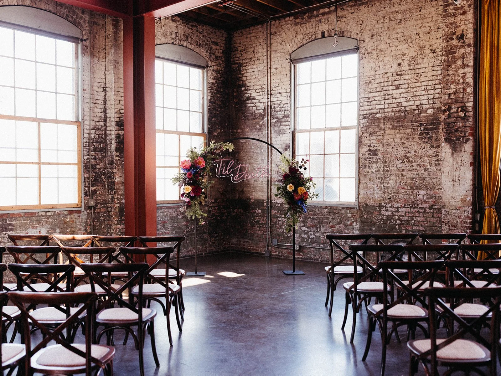 Industrial wedding ceremony setup featuring exposed brick walls, large windows, floral installations, and a neon "'Til Death" ceremony backdrop.