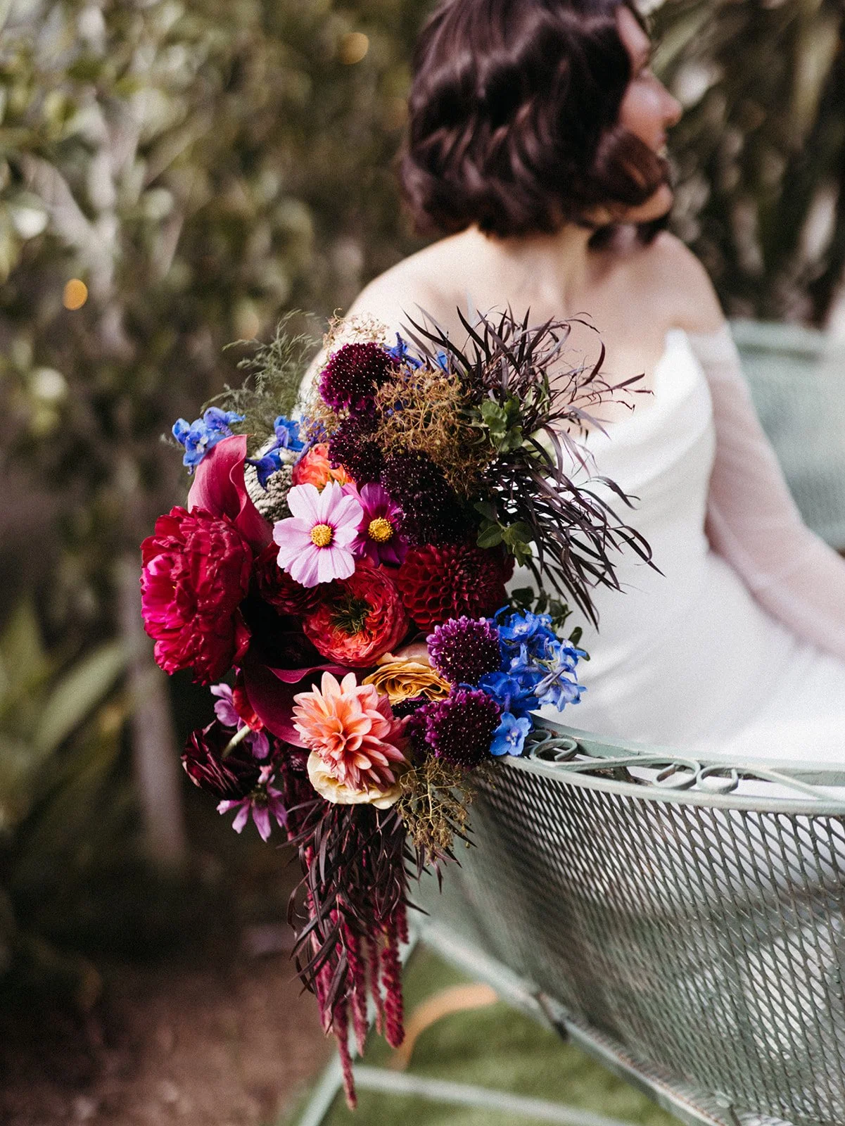 Close-up of a vibrant bridal bouquet featuring burgundy, pink, blue, and peach flowers resting on a vintage metal garden bench.