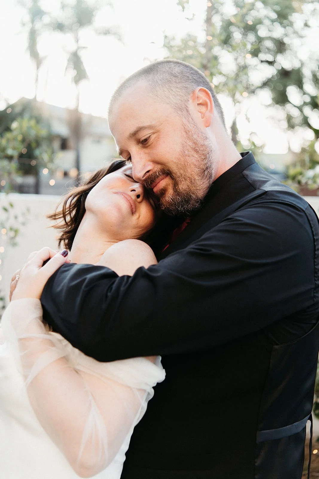 Bride and groom share a quiet embrace outdoors during golden hour, surrounded by greenery and soft twinkle lights after their wedding ceremony.