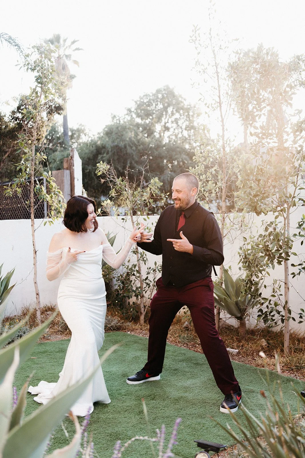 Bride and groom laugh and strike playful poses together during relaxed wedding portraits in a garden courtyard.