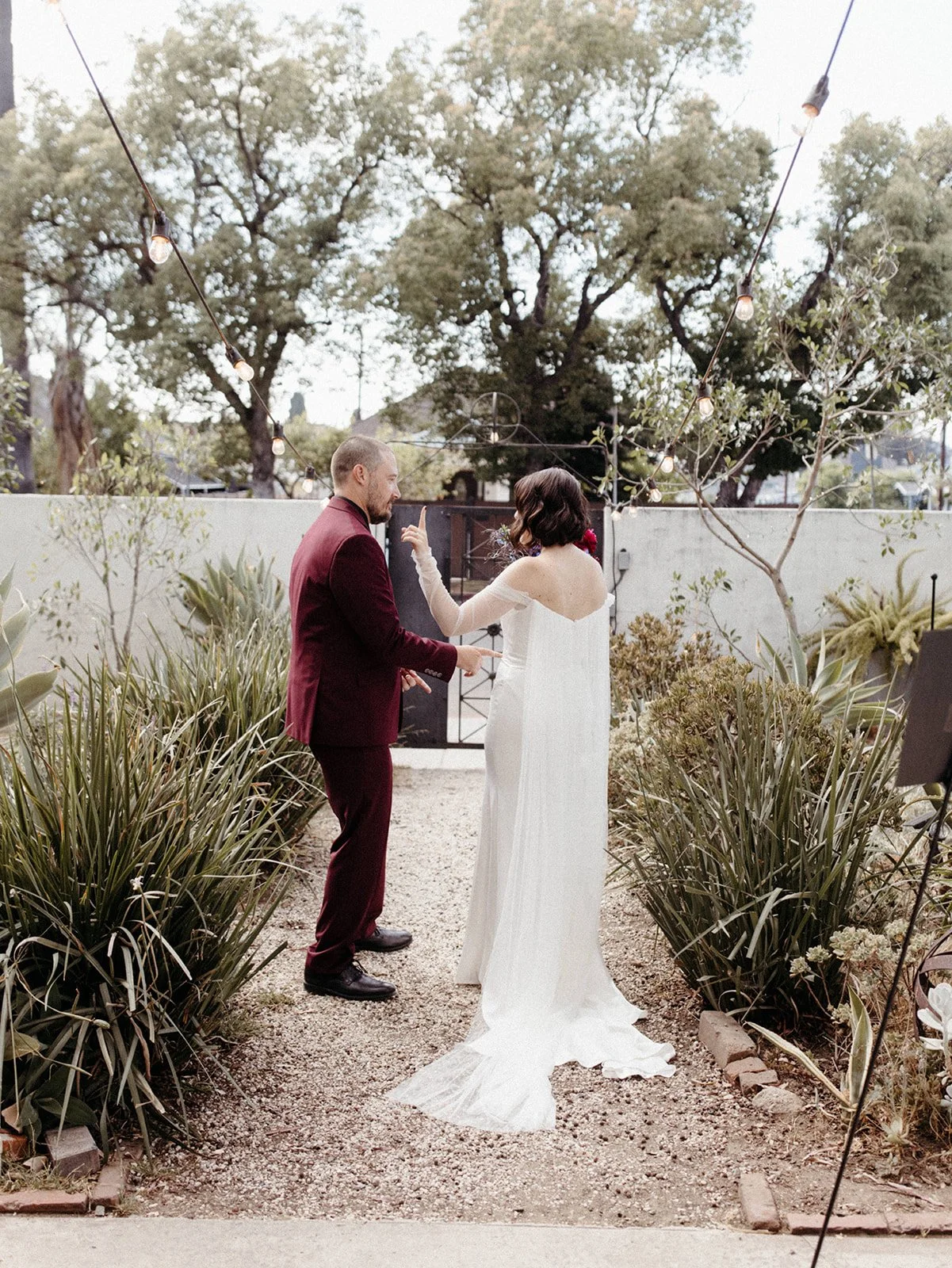 Bride and groom share a candid conversation in a garden courtyard during their wedding day first look, surrounded by greenery and string lights.