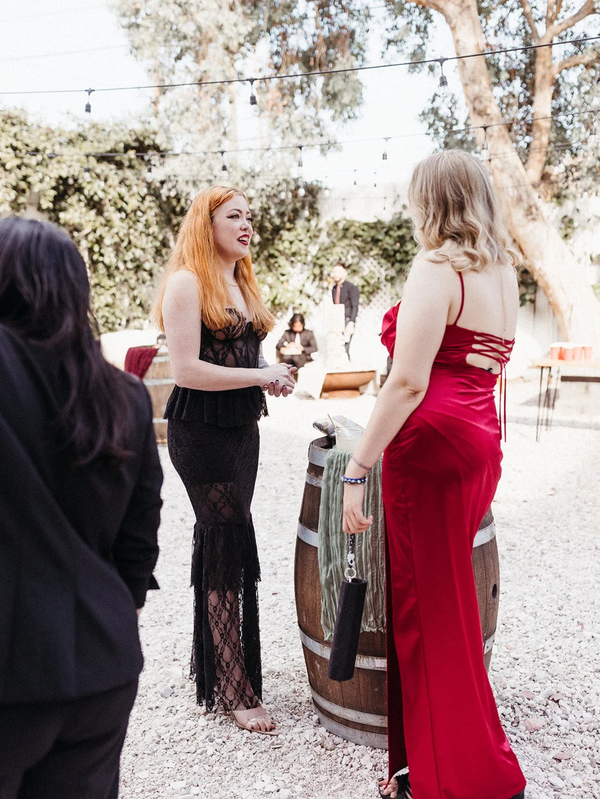 Wedding guests in black and red formal attire chat around cocktail tables during an outdoor cocktail hour reception.