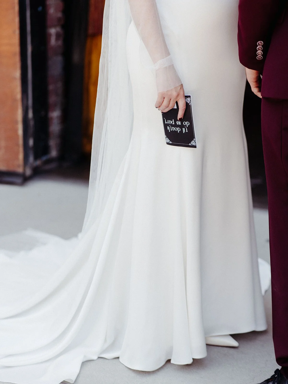 Bride in a modern white wedding gown holds a custom black vow book beside her groom during their private first look.