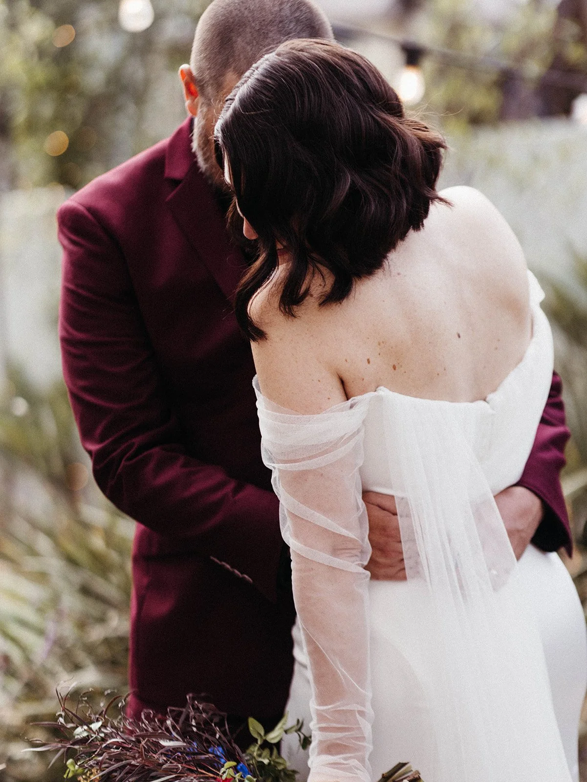 Intimate close-up of a bride and groom embracing in a garden setting, highlighting the bride's off-the-shoulder gown and flowing tulle sleeves.