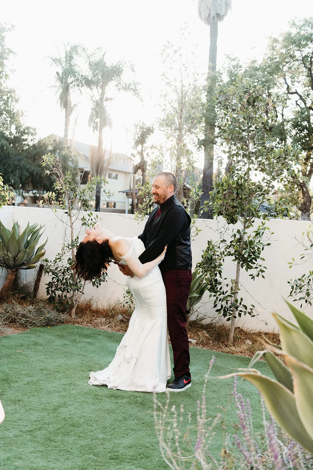 Groom dips the bride backward during a playful wedding portrait surrounded by palm trees and garden greenery.
