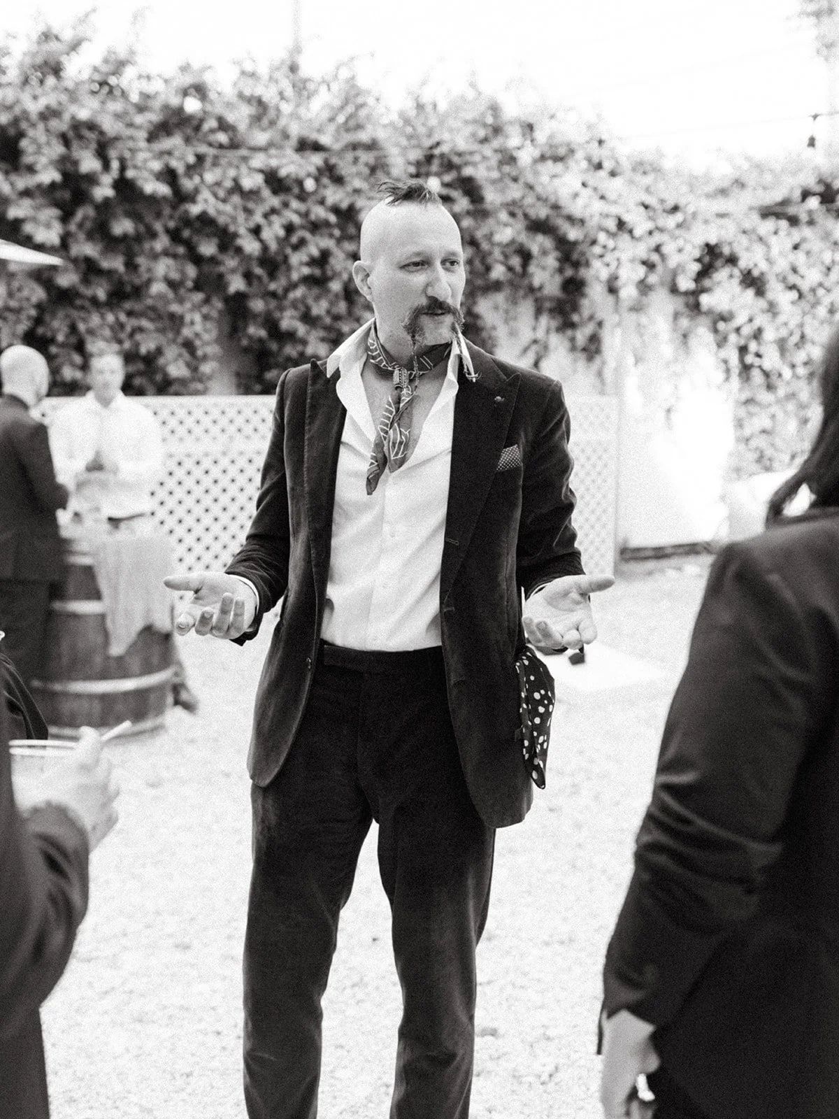 Wedding guest with a mohawk hairstyle, velvet blazer, and patterned neck scarf mingles during an alternative-inspired wedding cocktail hour.