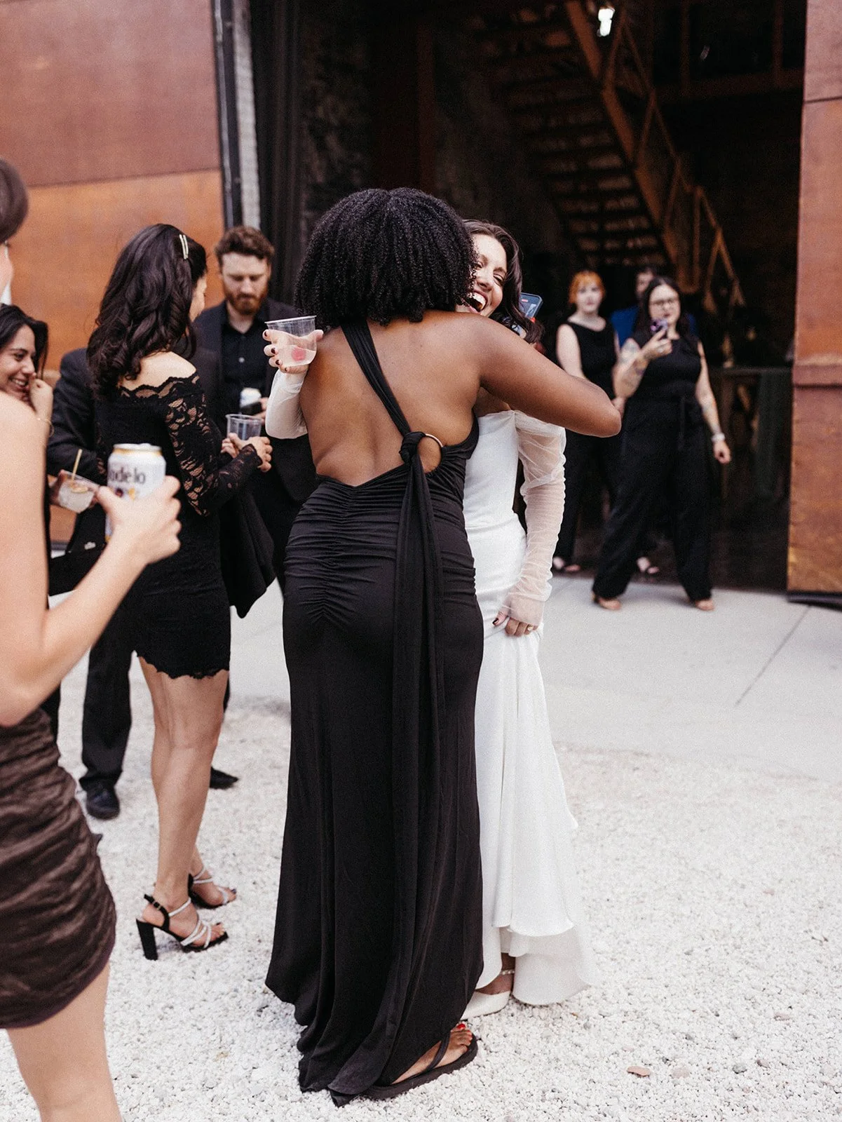 Bride in an off-the-shoulder wedding gown hugs a guest during cocktail hour outside an industrial wedding venue while friends mingle nearby.