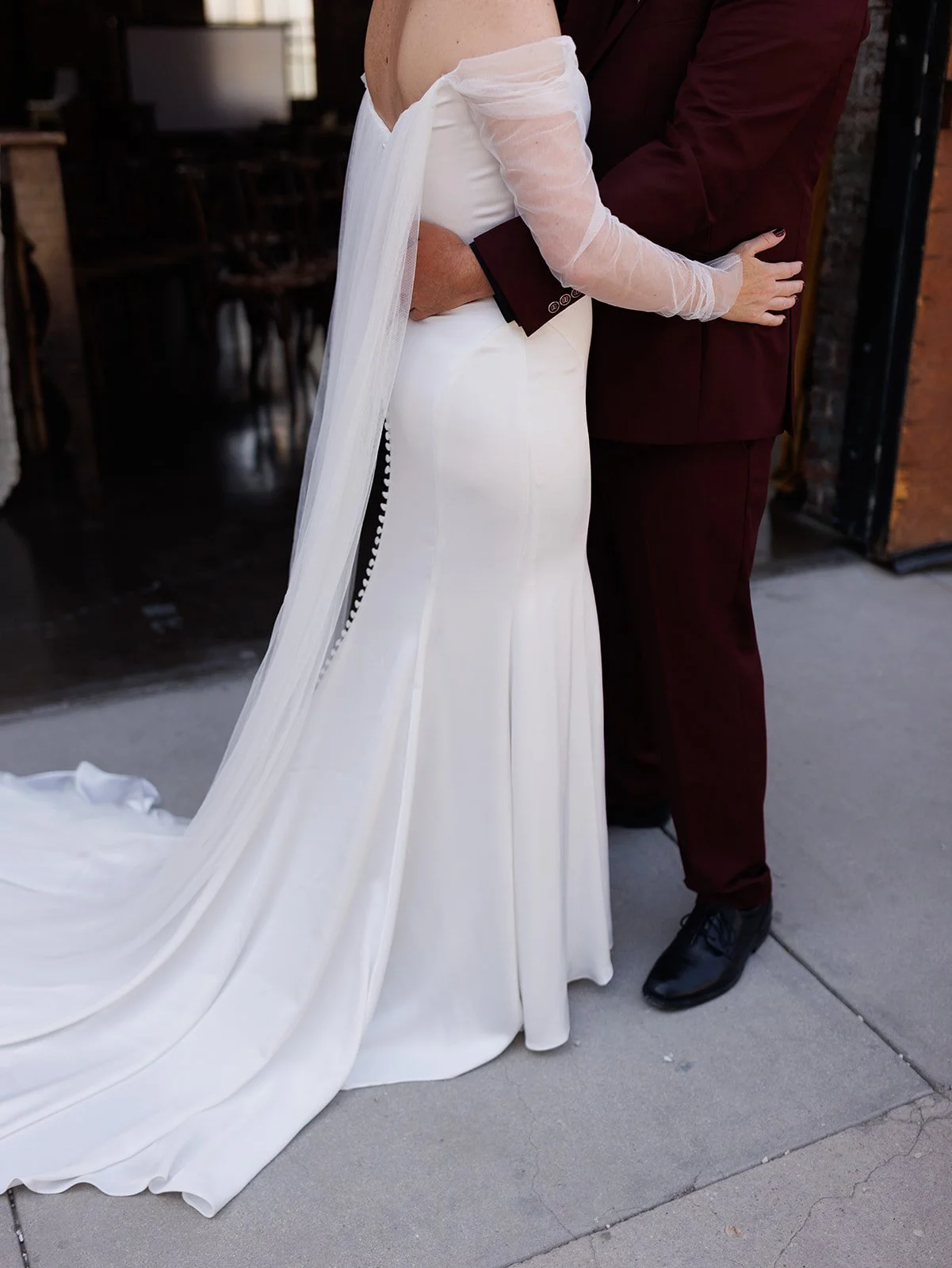 Bride in a fitted off-the-shoulder wedding gown with a flowing cape veil embraces groom in a burgundy suit outside an industrial wedding venue.