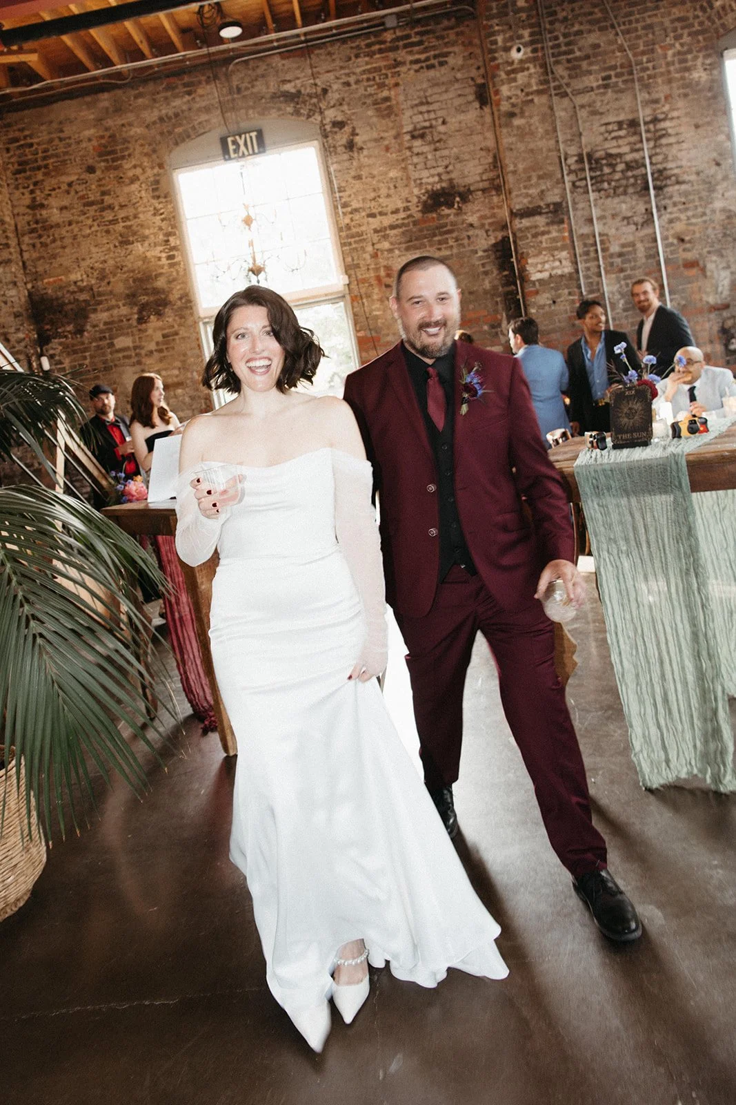 Newlyweds walk into their wedding reception smiling and holding drinks inside a historic industrial venue surrounded by guests and reception décor.