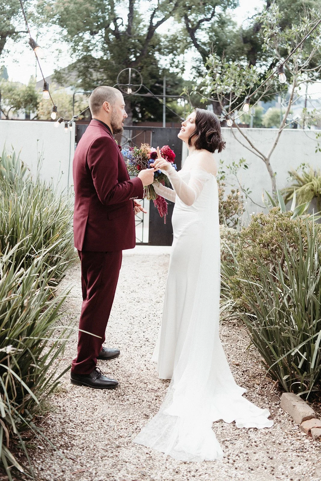 Bride and groom share a candid conversation in a garden courtyard during their wedding day first look, surrounded by greenery and string lights.