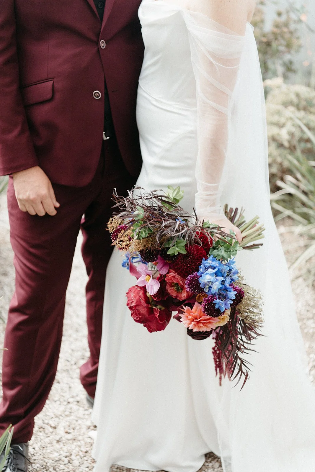 Close-up of a bride holding a vibrant wedding bouquet featuring burgundy, blue, pink, and coral blooms beside a groom in a burgundy suit.