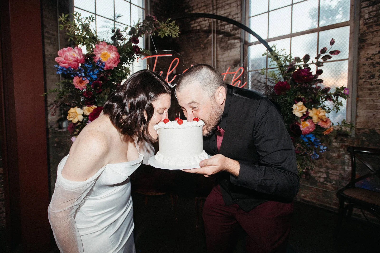 Bride and groom playfully taking bites of their wedding cake during the reception, surrounded by colorful florals and personalized design details at an unconventional wedding celebration.