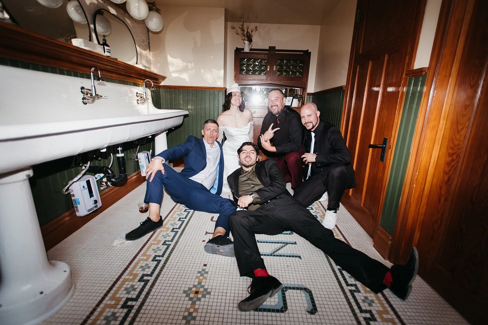Bride, groom, and wedding party posing for a playful group photo in a historic venue restroom during the wedding reception.