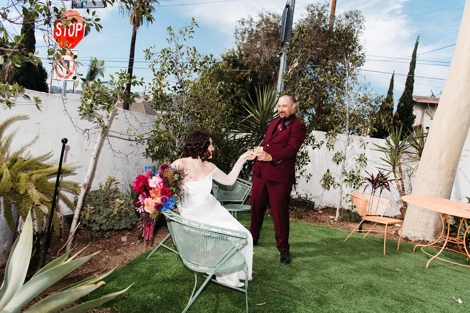 Bride and groom share a celebratory cocktail toast in a garden courtyard with colorful florals, vintage patio furniture, and lush greenery.
