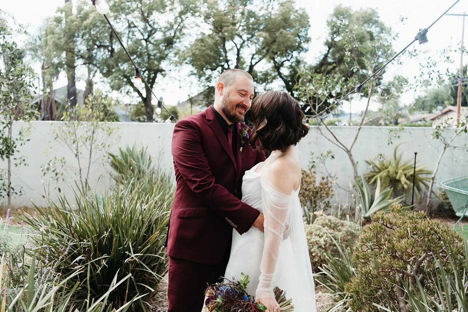 Bride and groom embrace in a garden setting beneath string lights, surrounded by desert-inspired landscaping and colorful wedding florals.