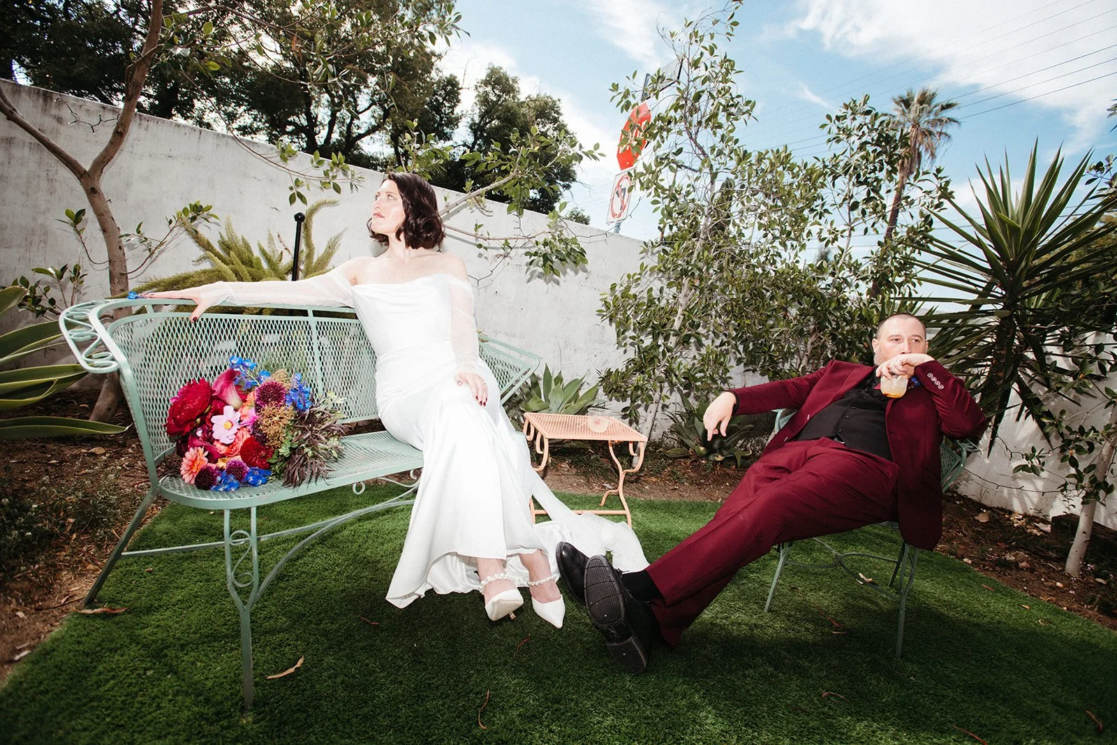 Bride and groom relax in a colorful outdoor lounge area with vintage furniture, cocktails, and vibrant floral arrangements during a modern wedding portrait session.