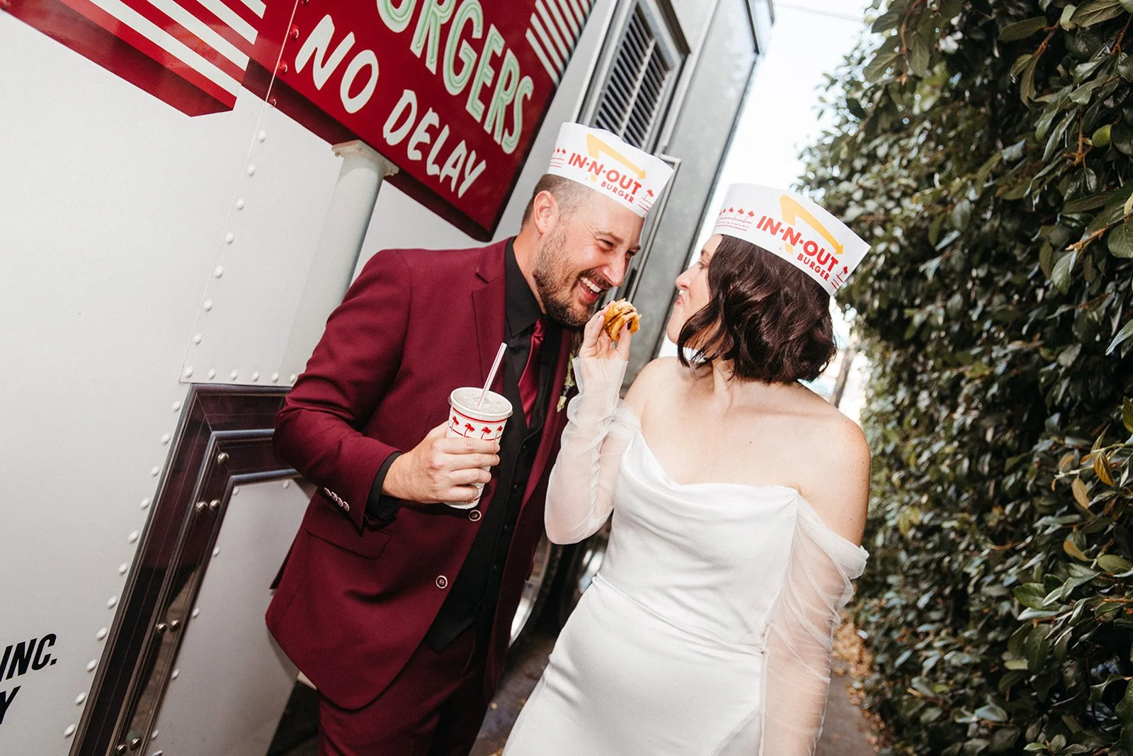 Newlywed couple wearing In-N-Out paper hats share burgers and milkshakes beside an In-N-Out food truck during their wedding celebration.