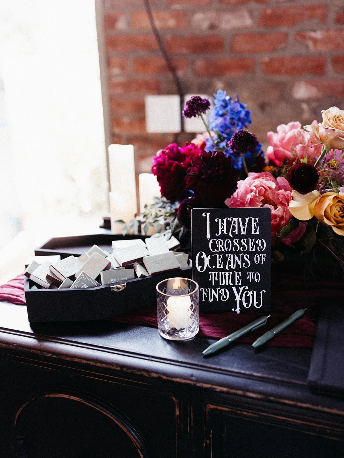 Travel-inspired wedding guest book display featuring colorful floral arrangements, candles, matchbooks, pens, and a sign reading “I Have Crossed Oceans of Time to Find You.”