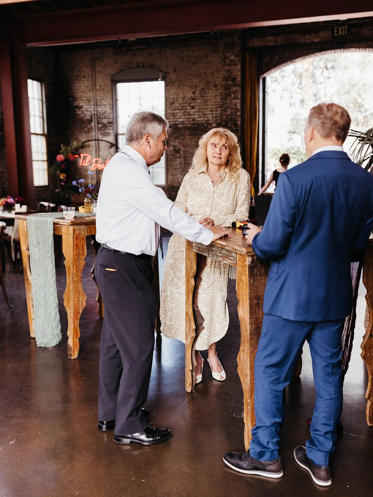 Wedding guests gather around rustic high-top tables during cocktail hour inside an industrial loft venue with exposed brick walls and warm ambient lighting.
