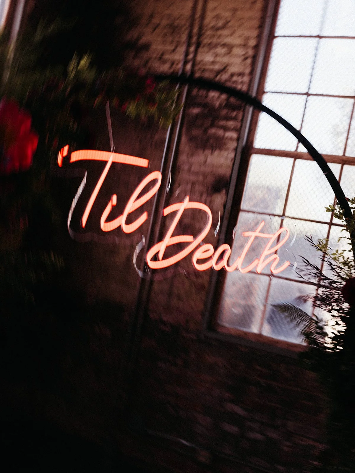 Close-up of a glowing “Til Death” neon sign framed by lush burgundy, pink, cream, and blue floral arrangements, creating a bold and romantic focal point for a modern wedding ceremony backdrop.