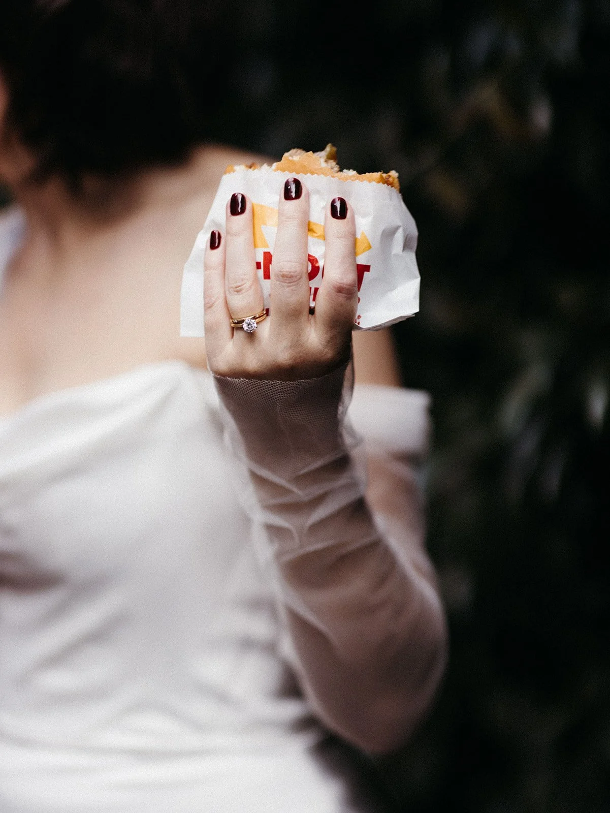 Close-up of the bride’s hand holding an In-N-Out burger during the wedding reception, showcasing her engagement ring and dark burgundy manicure.