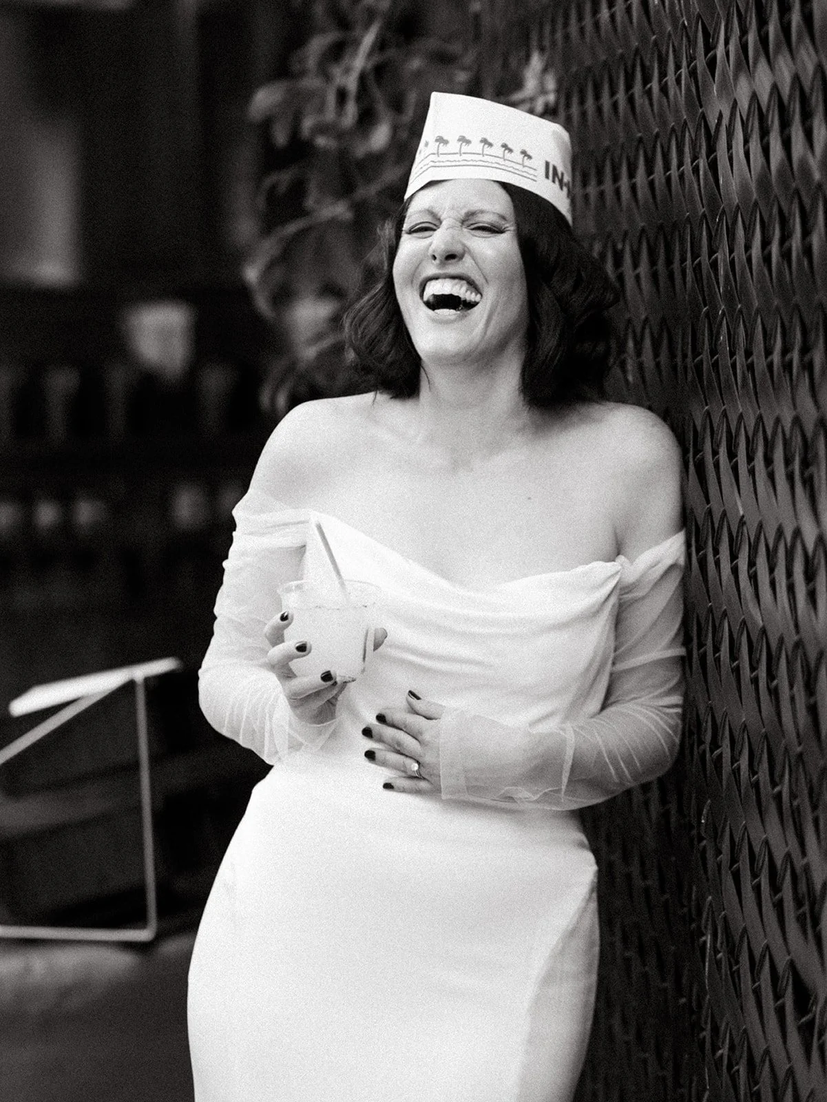 Bride in an off-the-shoulder wedding gown laughs while holding a drink during the reception, wearing a paper In-N-Out hat in a playful candid moment.