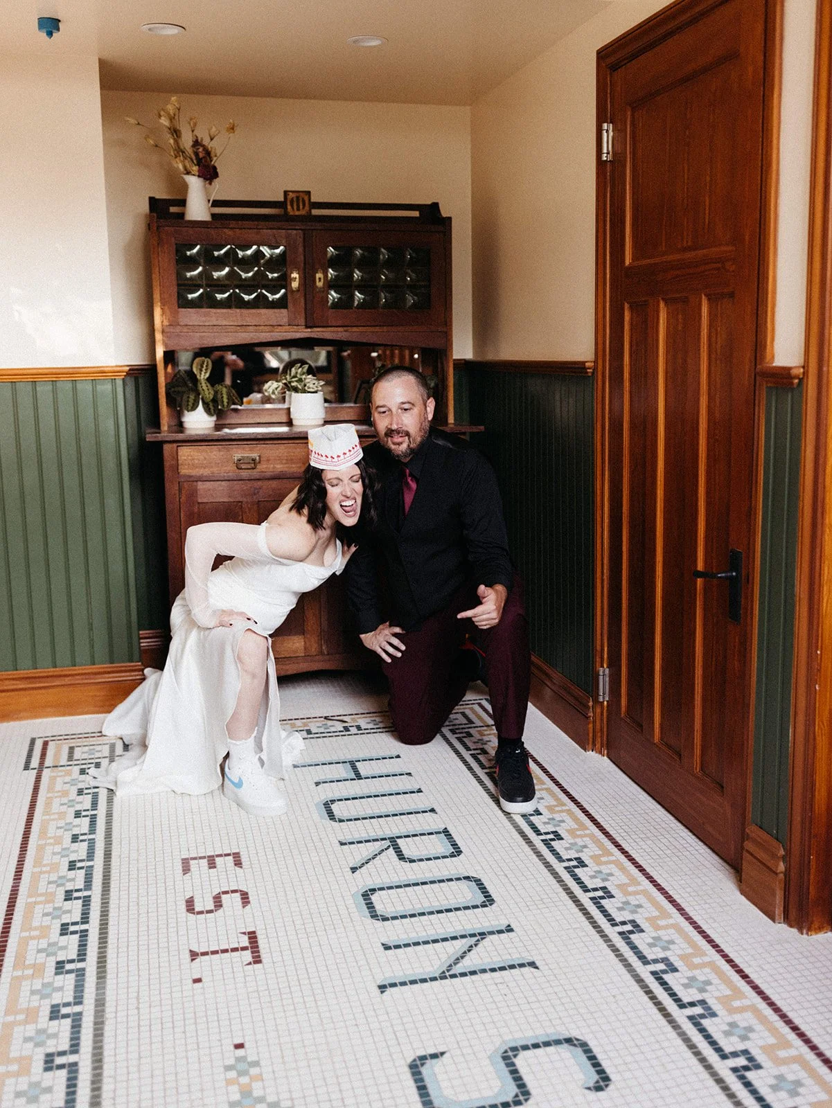 Newlywed couple poses playfully with In-N-Out burgers while wearing wedding attire and paper hats inside a historic building during their wedding after-party celebration.