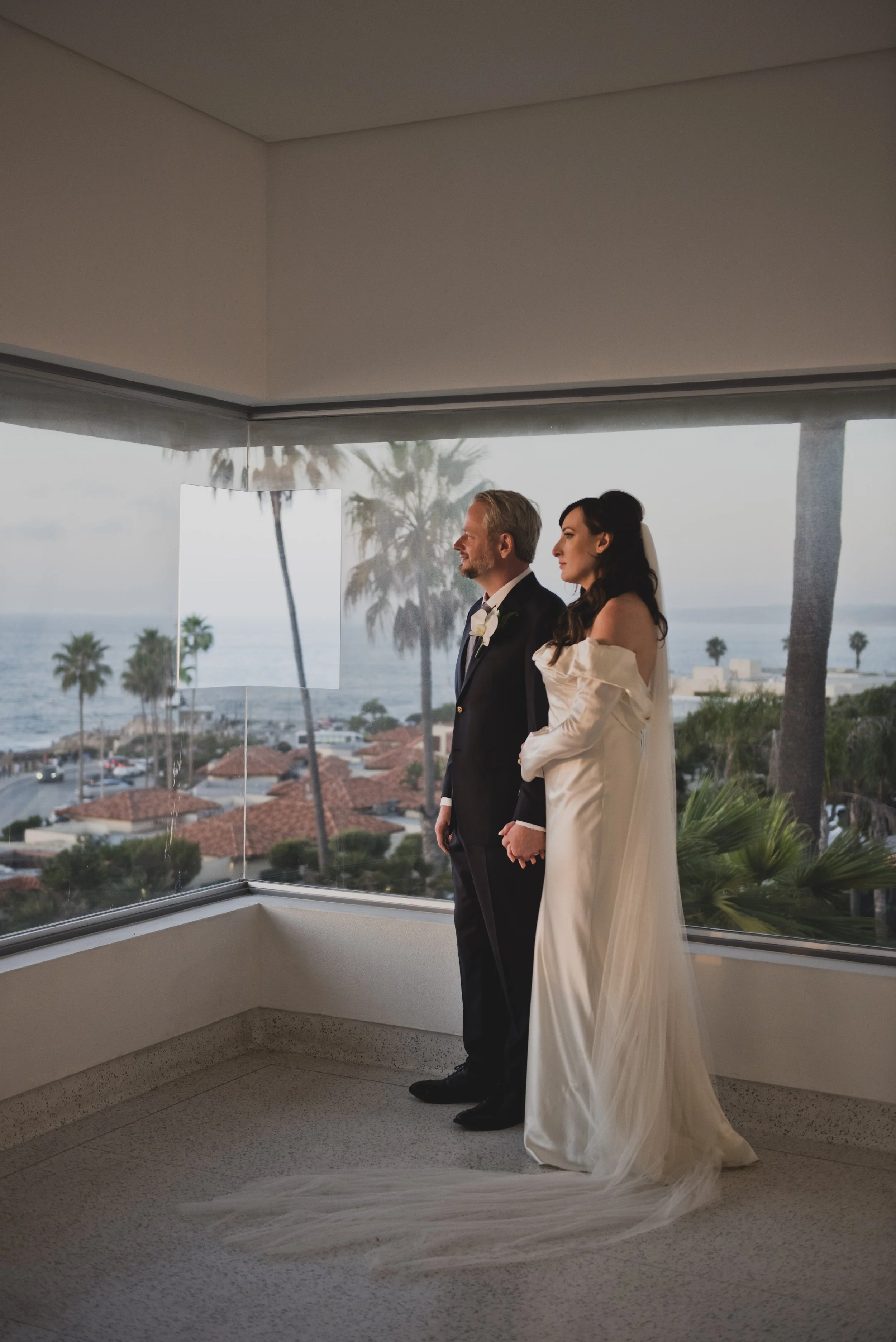 Bride and groom embracing in front of floor-to-ceiling windows overlooking the ocean, creating a romantic museum wedding portrait at sunset.