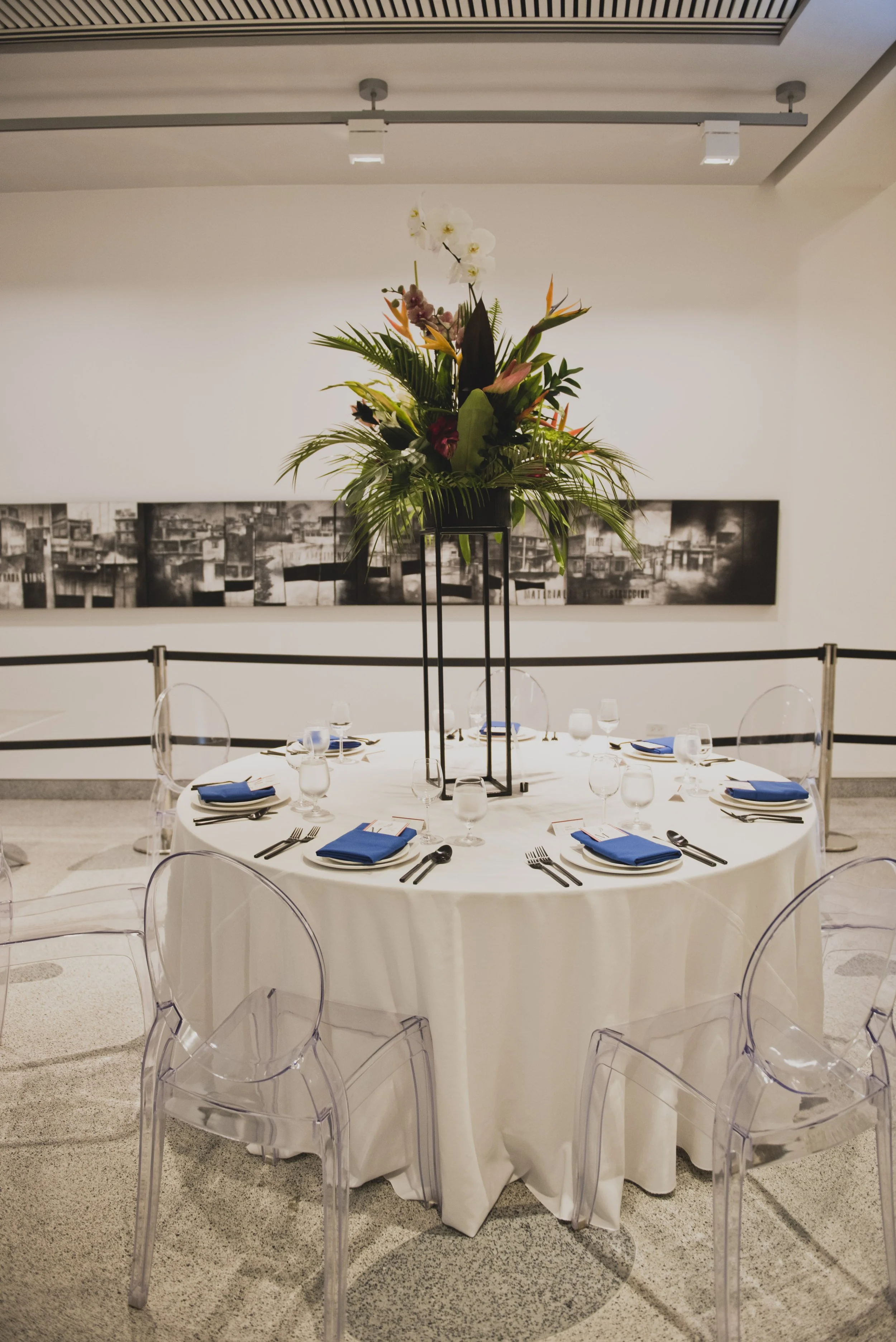 Elegant wedding reception table with a tall tropical floral centerpiece, clear ghost chairs, blue napkins, and modern place settings inside a contemporary art museum.