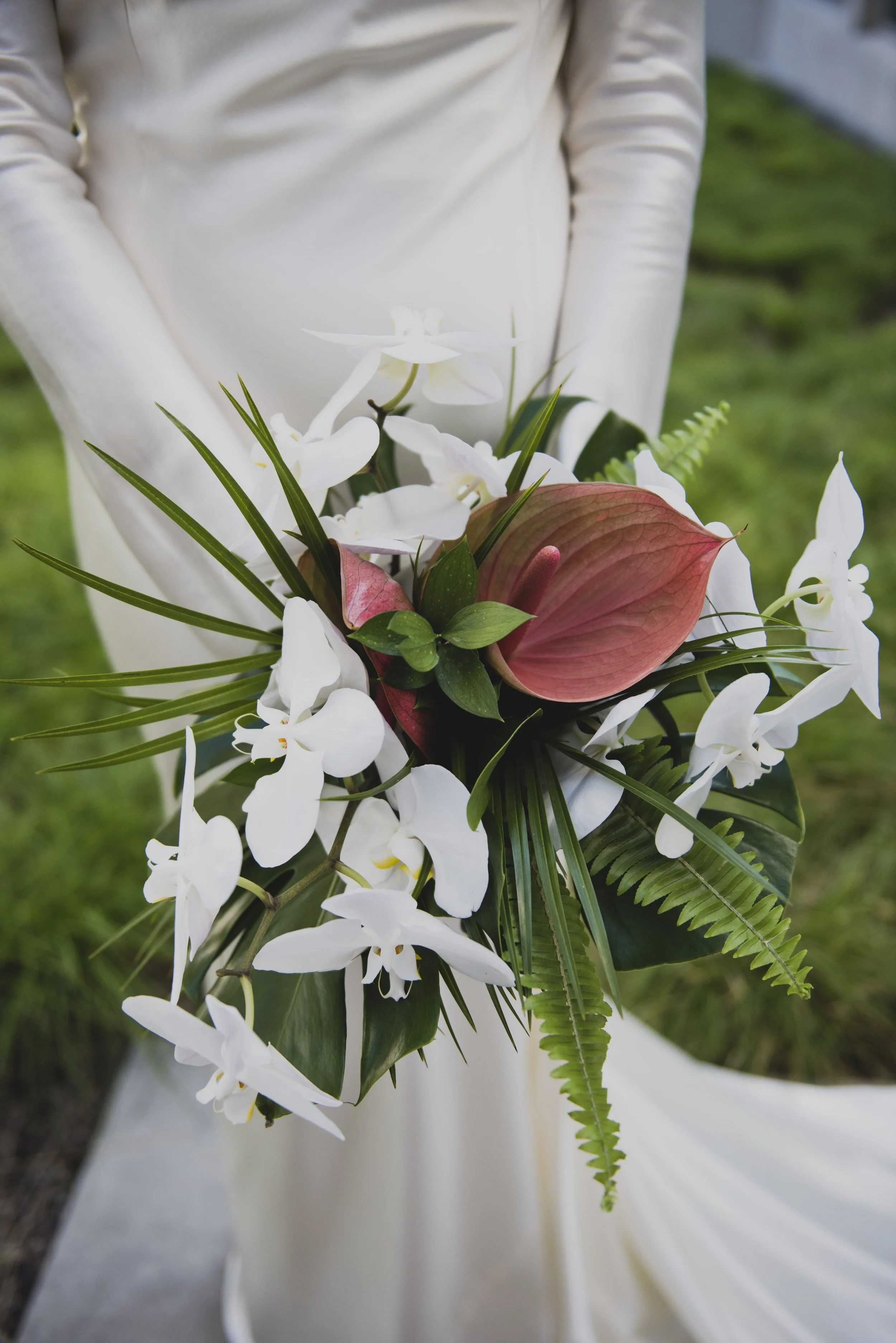 Bride holding a modern tropical bridal bouquet featuring white orchids, blush anthuriums, lush greenery, and palm foliage against her elegant wedding gown.