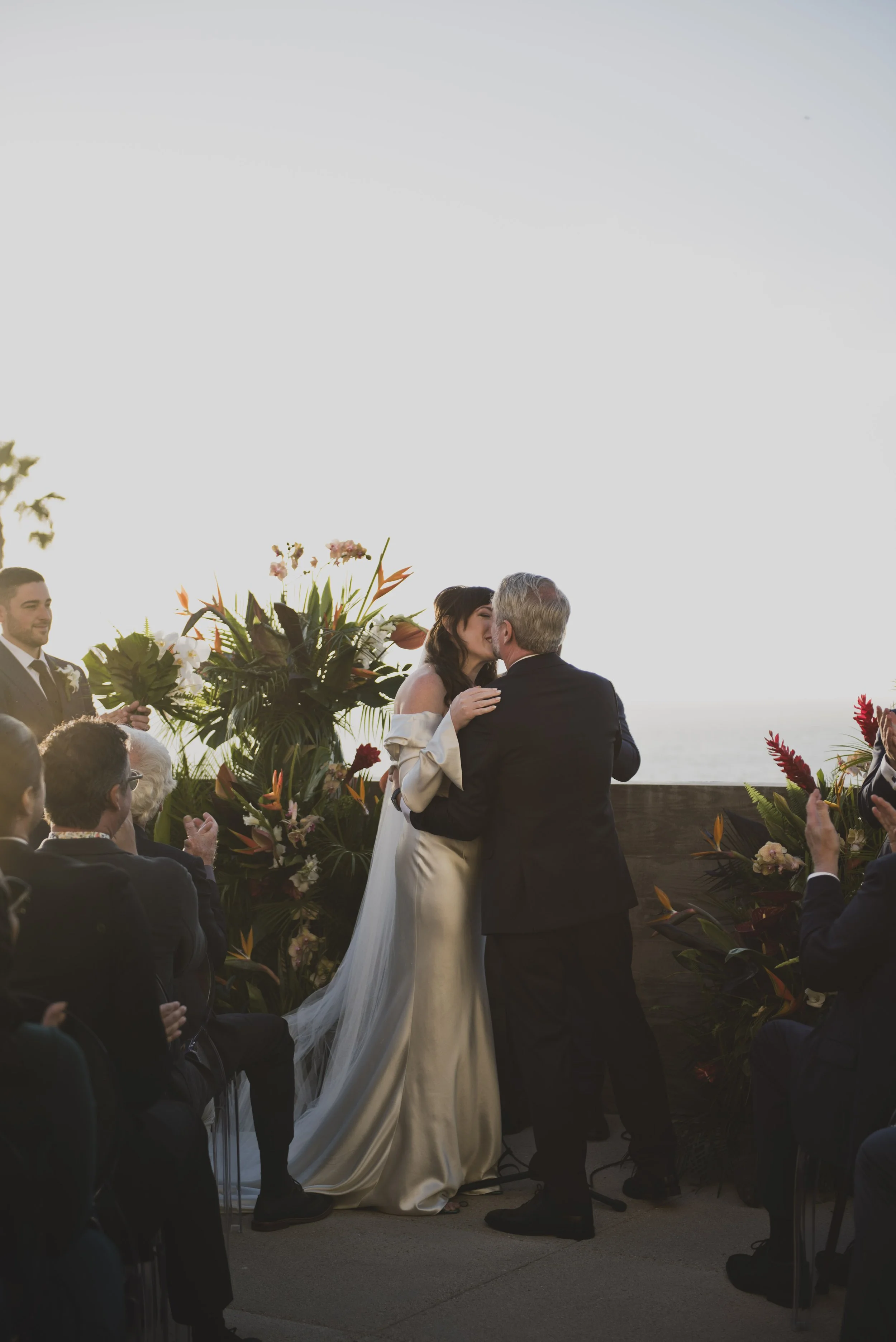 Groom embracing the bride during an emotional moment at an outdoor oceanfront wedding ceremony at sunset.