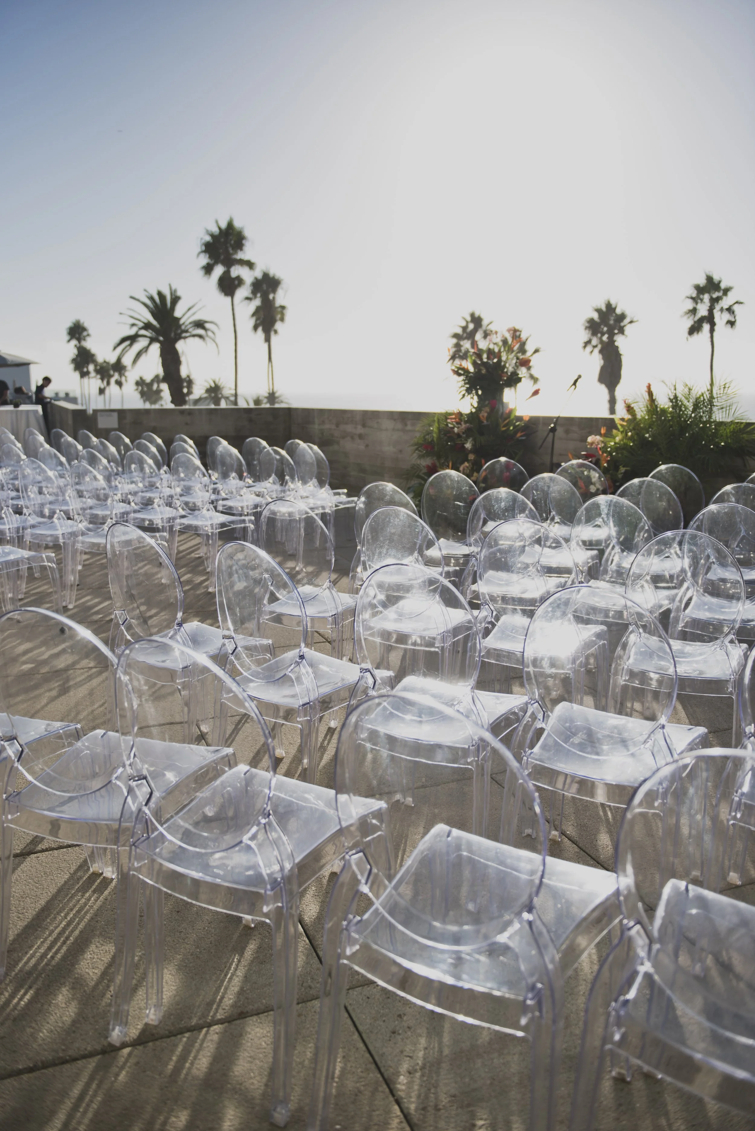 Clear ghost chairs arranged for an oceanfront wedding ceremony with tropical floral arrangements, palm trees, and panoramic coastal views.