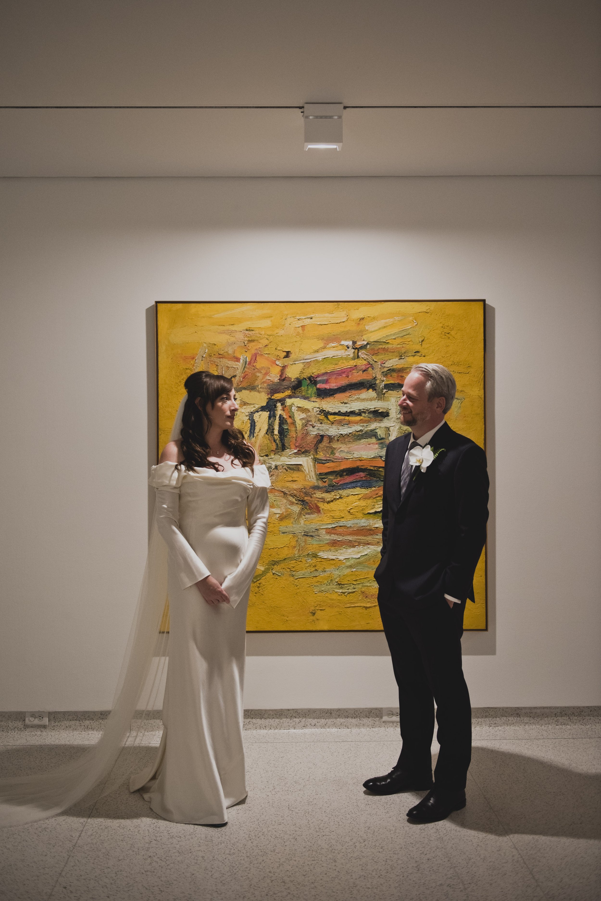 Bride and groom standing in front of a vibrant abstract painting inside a modern art museum, sharing a quiet moment before their wedding ceremony.