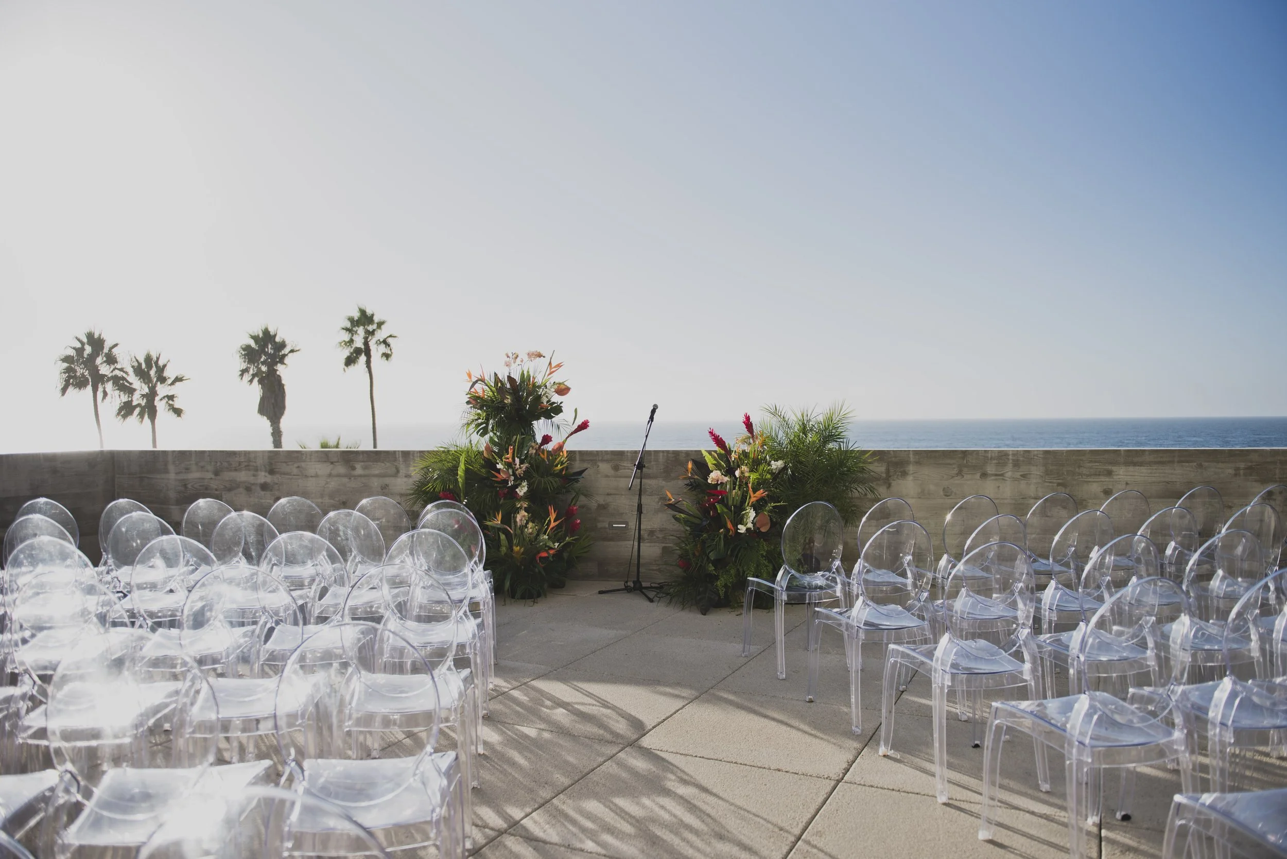 Clear chairs set up during intimate oceanfront wedding ceremony with tropical floral arrangements 