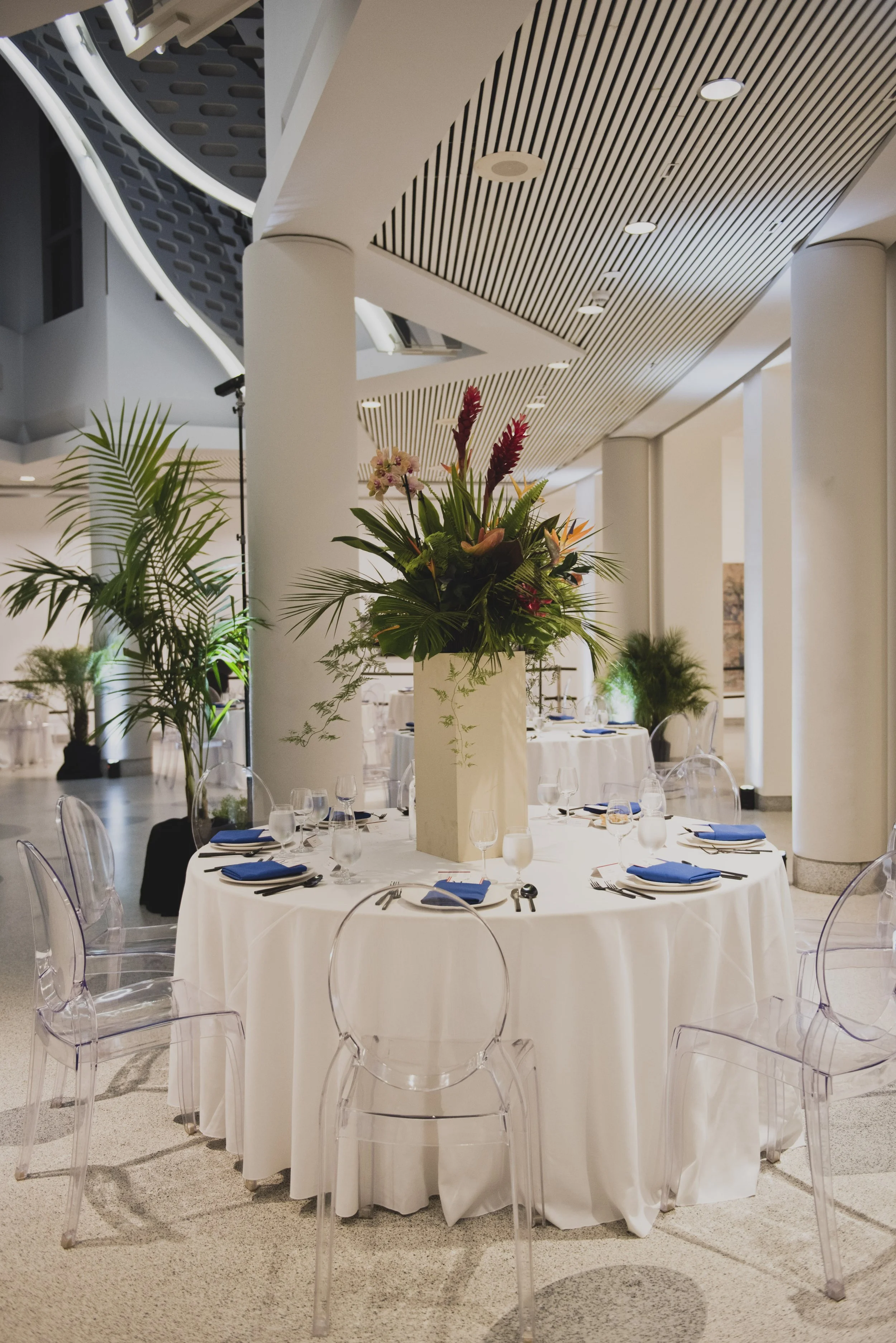Modern wedding reception table with a tropical floral centerpiece, clear ghost chairs, crisp white linens, and elegant place settings inside a contemporary museum venue.
