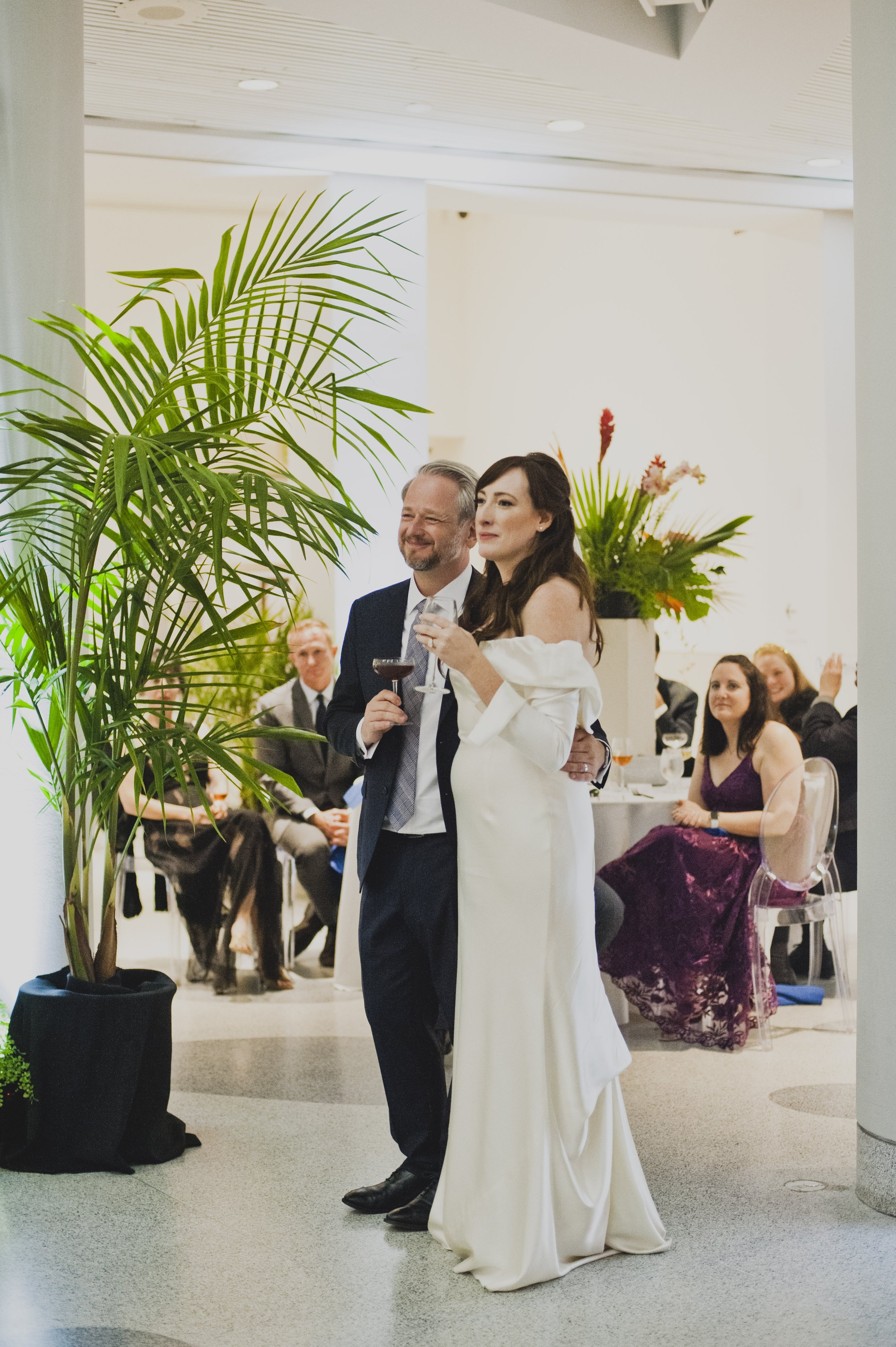 Newly married couple holding wine glasses and listening to wedding toasts while surrounded by family and friends at their modern museum wedding reception.