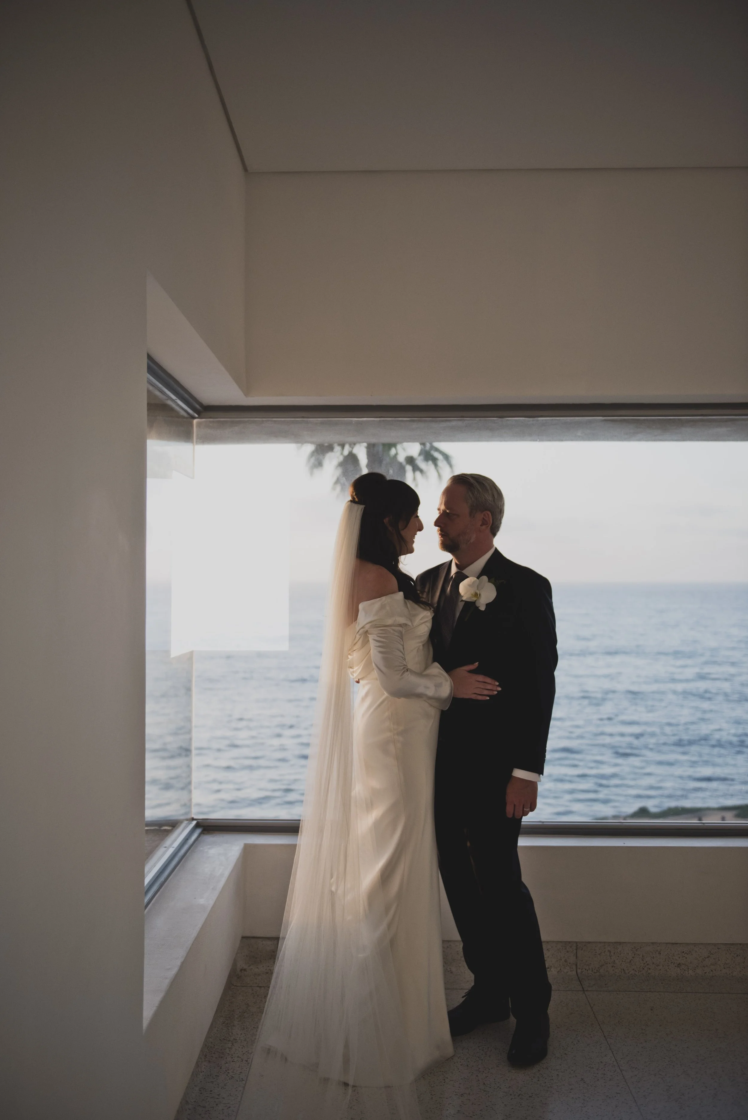 Bride and groom standing across from each other in front of expansive ocean-view windows inside a modern museum during golden hour.
