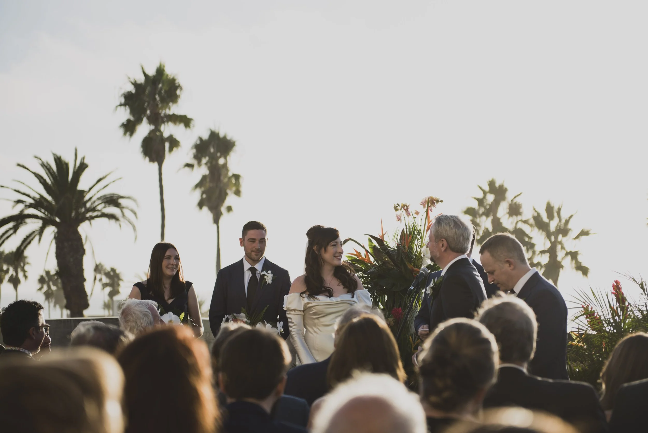Bride and groom exchanging vows during an intimate oceanfront wedding ceremony with tropical floral arrangements and guests seated outdoors.