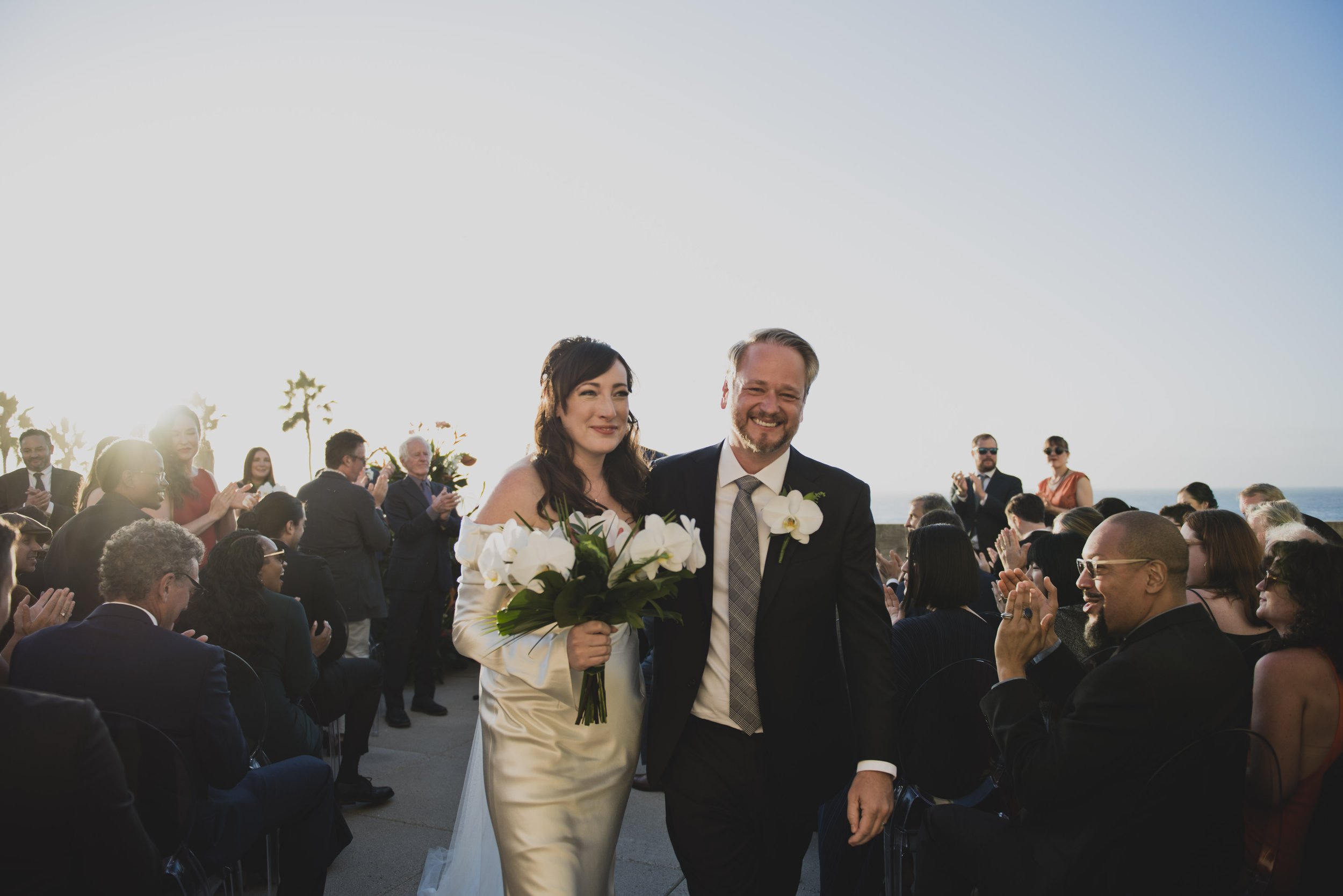 Newly married couple walking down the aisle after their oceanfront wedding ceremony while guests applaud and celebrate around them.